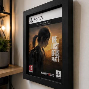 Puede incluir: Un marco de pared negro que muestra la portada del videojuego The Last of Us Part I para PS5. La ilustración presenta a un personaje en un entorno postapocalíptico oscuro con el título del juego, el logo de Naughty Dog y una etiqueta de 18.