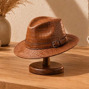 Peut inclure: Un chapeau fedora marron avec un motif texturé en peau de crocodile, une calotte pincée et une bande assortie dotée d'une boucle en métal argenté. Le chapeau est présenté sur un support en bois circulaire, mettant en valeur son bord structuré.