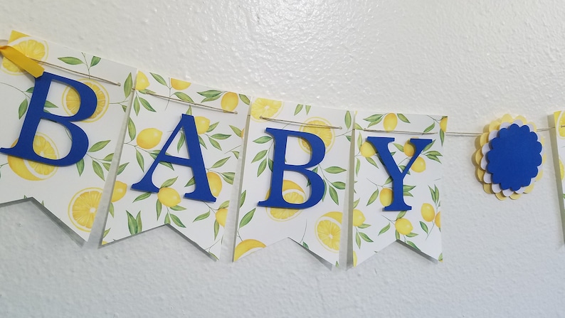 May include: A blue and yellow baby shower banner with the word "BABY" in blue letters. The banner is decorated with a lemon print.