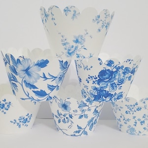 May include: Six white cupcake liners with a scalloped edge are arranged in a pyramid. Each liner is decorated with a blue floral pattern. The floral designs vary, featuring roses, hydrangeas, and other flowers.