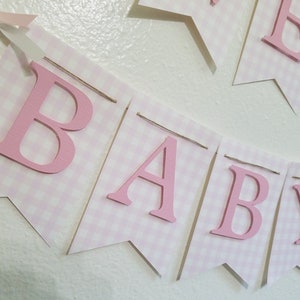 以下が含まれることがあります： ピンクと白のギンガムチェックのバナーで、ピンクの文字で「BABY」と書かれています。バナーは紐でつながれており、上部にピンクのリボンが付いています。ベビーシャワーや保育園の装飾に最適です。