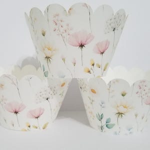 Puede incluir: Tres envoltorios blancos para cupcakes con bordes festoneados. Cada envoltorio está decorado con un diseño floral de acuarela en tonos rosa pálido, amarillo y verde. Las flores incluyen margaritas y otras flores silvestres.