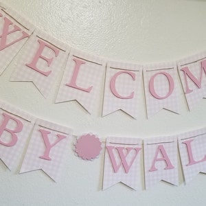 以下が含まれることがあります： ピンク色の文字で「WELCOME BABY WALIOR」と書かれたバナー。白いペナントにはピンクのギンガムチェック模様が施されています。言葉の間にはピンク色のスカラップサークルがあります。