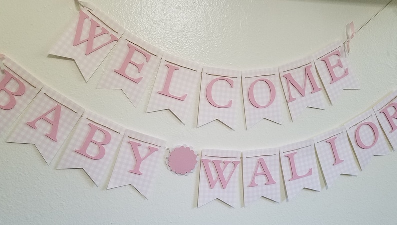 以下が含まれることがあります： ピンク色の文字で「WELCOME BABY WALIOR」と書かれたバナー。白いペナントにはピンクのギンガムチェック模様が施されています。言葉の間にはピンク色のスカラップサークルがあります。