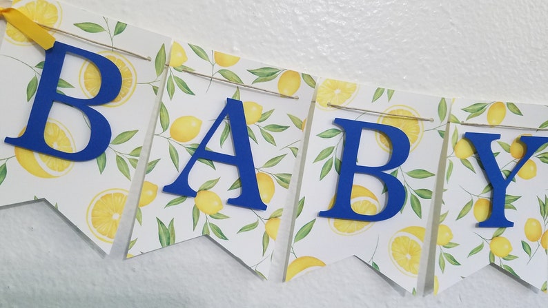 May include: A blue and white baby shower banner with the word "BABY" in blue letters on a white background with a lemon pattern.