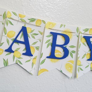 May include: A blue and yellow baby shower banner with the word "BABY" in blue letters on a white background with a lemon pattern.