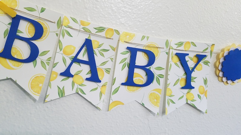 May include: A blue and yellow baby shower banner with the word "BABY" in blue letters on a white background with a lemon pattern.