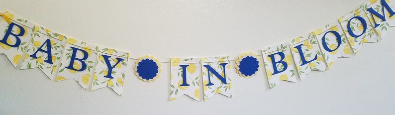 May include: A baby shower banner with a lemon print background and blue letters that read "Baby in Bloom".