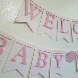 以下が含まれることがあります： ピンクと白のギンガムチェックのバナーで、ピンクの文字で「WELCOME BABY」と書かれています。バナーは紐でつながっており、ベビーシャワーや保育園の装飾用である可能性があります。