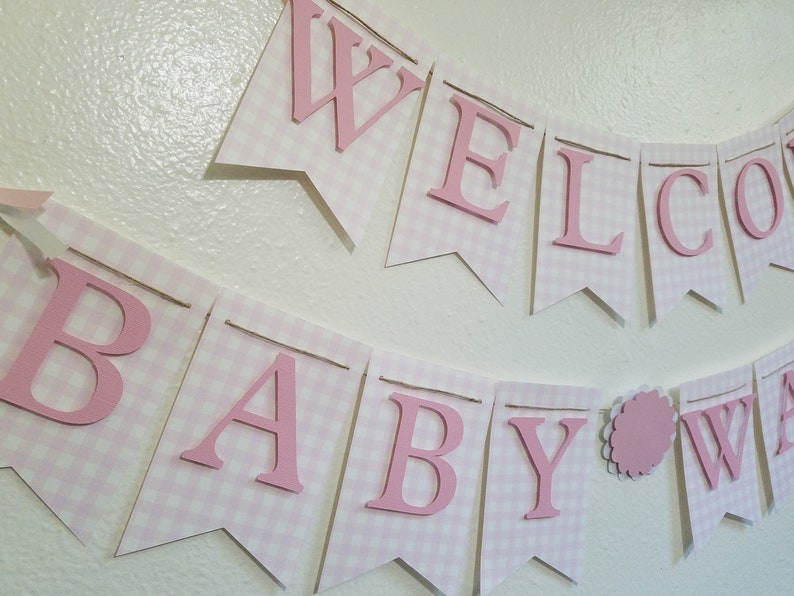 以下が含まれることがあります： ピンクと白のギンガムチェックのバナーで、ピンクの文字で「WELCOME BABY」と書かれています。バナーは紐でつながっており、ベビーシャワーや保育園の装飾用である可能性があります。