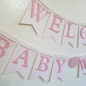 以下が含まれることがあります： 「WELCOME BABY WA」と書かれたピンクと白のギンガムチェックのバナー。文字はピンク色で盛り上がっており、個々のペナントフラッグに取り付けられています。バナーはひもでつながれており、ベビーシャワー用の装飾品です。