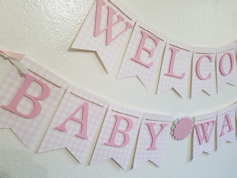 以下が含まれることがあります： 「WELCOME BABY WA」と書かれたピンクと白のギンガムチェックのバナー。文字はピンク色で盛り上がっており、個々のペナントフラッグに取り付けられています。バナーはひもでつながれており、ベビーシャワー用の装飾品です。