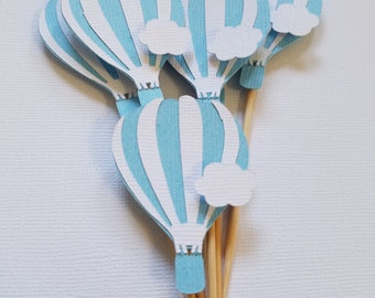 May include: A collection of paper hot air balloon cutouts in light blue and white stripes, each featuring a small white cloud accent. These decorative paper shapes are attached to thin wooden sticks, creating a set of toppers for display.