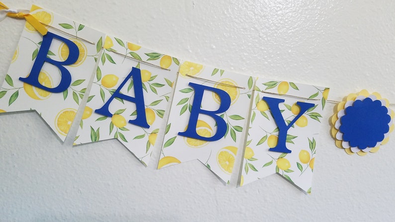 May include: A blue and yellow baby shower banner with the word "BABY" in blue letters. The banner is decorated with a lemon print.