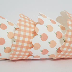 May include: Three cupcake liners with a peach and gingham pattern. The liners are white with peach-colored gingham and peach fruit designs.
