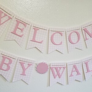 以下が含まれることがあります： ピンク色の文字で「WELCOME BABY WALLIOR」と書かれたバナー。バナーは、ピンクと白のギンガムチェック模様の白いペナントでできています。ピンク色のスカラップサークルが単語の間にあります。