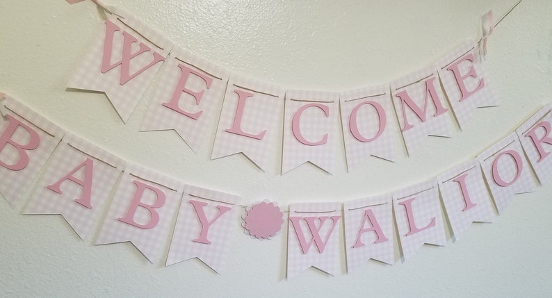 以下が含まれることがあります： ピンク色の文字で「WELCOME BABY WALLIOR」と書かれたバナー。バナーは、ピンクと白のギンガムチェック模様の白いペナントでできています。ピンク色のスカラップサークルが単語の間にあります。