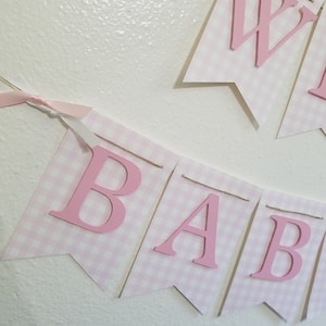 以下が含まれることがあります： 「BABY」と「WELCOME」の文字が入ったピンクと白のギンガムチェックのバナー。文字は濃いピンク色で、ひもでバナーに固定されています。ピンクのリボンがバナーに結ばれています。バナーは白い壁に掛けられています。