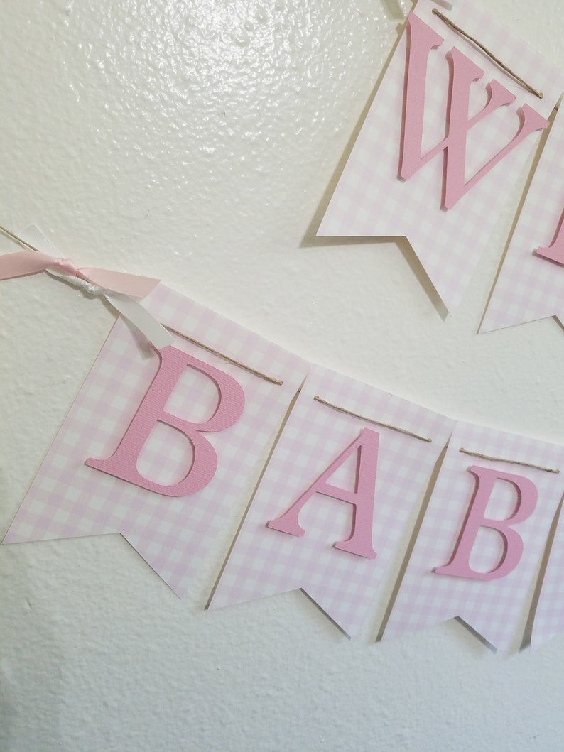 以下が含まれることがあります： 「BABY」と「WELCOME」の文字が入ったピンクと白のギンガムチェックのバナー。文字は濃いピンク色で、ひもでバナーに固定されています。ピンクのリボンがバナーに結ばれています。バナーは白い壁に掛けられています。