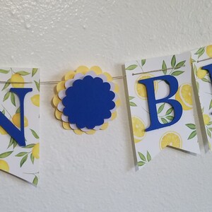 May include: A yellow and white banner with the words "In Bloom" in blue letters. The banner is decorated with a lemon print.