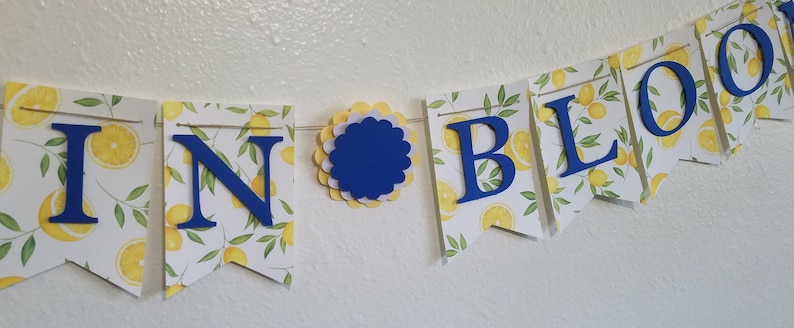 May include: A yellow and white banner with the words "In Bloom" in blue letters. The banner is decorated with a lemon print.