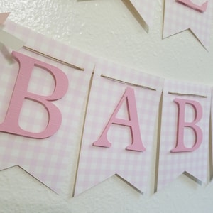 以下が含まれることがあります： 「BABY」の文字が書かれたピンクと白のギンガムチェックのバナー。文字はピンク色で、個々のペナントフラッグに取り付けられています。バナーは紐でつながれており、ピンクのリボンがアクセントになっており、ベビーシャワーや保育園の装飾に最適です。