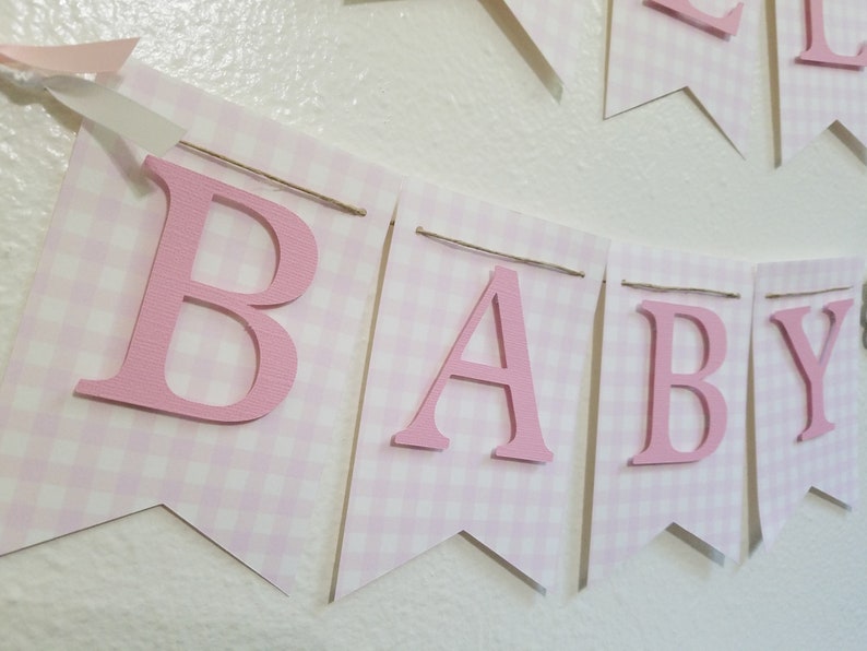 以下が含まれることがあります： 「BABY」の文字が書かれたピンクと白のギンガムチェックのバナー。文字はピンク色で、個々のペナントフラッグに取り付けられています。バナーは紐でつながれており、ピンクのリボンがアクセントになっており、ベビーシャワーや保育園の装飾に最適です。