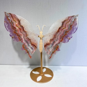 Puede incluir: Una escultura decorativa de mariposa con dos grandes alas translúcidas hechas de ágata bandeada en tonos morado, blanco y naranja. El cuerpo de la mariposa está elaborado en metal dorado y montado sobre un soporte de metal dorado a juego.