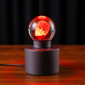 Może przedstawiać: Lampa stołowa z przezroczystą szklaną kulą, wewnątrz której znajduje się świecąca na czerwono ludzka czaszka. Kula spoczywa na cylindrycznej, matowej czarnej podstawie. Czarny przewód zasilający wychodzi z podstawy, emitując czerwone światło.