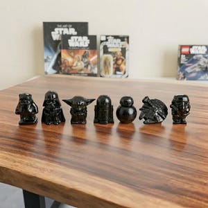 Può includere: Un set di sette piccole statuette nere e lucide dei personaggi di Star Wars disposte in linea orizzontale. Le figure includono un Ewok, Darth Vader, Yoda, R2-D2, BB-8, il Millennium Falcon e uno Stormtrooper, tutte con una finitura liscia.