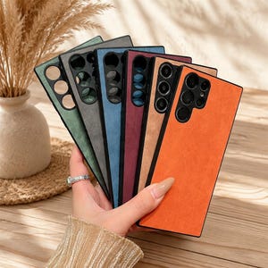 Peut inclure: Une collection d'étuis pour smartphone rectangulaires avec une finition texturée semblable à du daim, disposés en éventail. Les étuis sont disponibles en orange, beige, bordeaux, bleu, gris et vert, chacun avec un bloc photo noir.