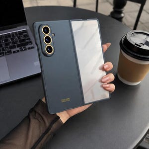 May include: A dark gray foldable smartphone case with a matte finish, featuring a vertical camera module with three gold-rimmed lenses and a glossy panel on the right side. The text FOLD CASE DESIGNED BY SKS is printed in gold at the bottom.