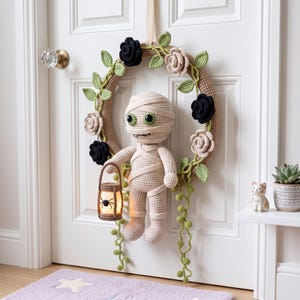 May include: A crochet wreath featuring a beige mummy figure holding a small lantern with a spider graphic. The wreath is decorated with black and beige crocheted roses and green vines with leaves, all suspended by a ribbon on a white door.