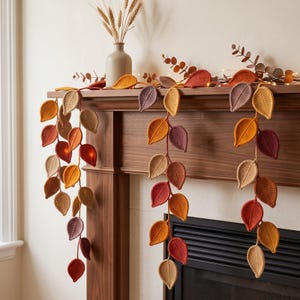 May include: A decorative garland featuring numerous crocheted leaves in shades of orange, yellow, brown, and purple. The leaves are arranged in clusters along a central cord, creating a cascading effect with varied textures and autumn-inspired colors.