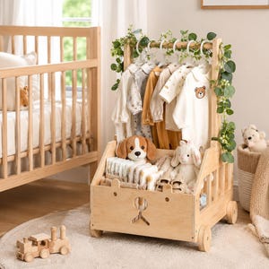 May include: A light-colored wooden rolling garment rack featuring a hanging rod with white plastic hangers holding baby clothes, a lower storage bin containing folded diapers and plush toys, and decorative green vines draped over the top frame.
