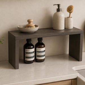 May include: A dark wood rectangular shelf riser with a flat top and two side supports. The shelf holds a small ceramic bowl with a wooden scrub brush, a beige soap dispenser, and a matching brush holder. Two amber glass bottles sit underneath.