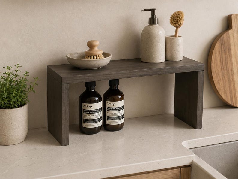 May include: A dark wood rectangular shelf riser with a flat top and two side supports. The shelf holds a small ceramic bowl with a wooden scrub brush, a beige soap dispenser, and a matching brush holder. Two amber glass bottles sit underneath.