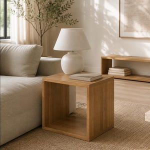 Puede incluir: Una mesa auxiliar cuadrada de madera clara con el centro abierto, rematada con una lámpara de mesa de cerámica blanca con pantalla de tela cónica y un libro rectangular colocado plano sobre la superficie junto a la base de la lámpara.