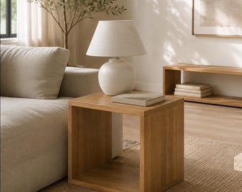 May include: A square, light-toned wooden side table with an open center, topped with a white ceramic table lamp featuring a tapered fabric shade and a rectangular book placed flat on the surface next to the lamp base.