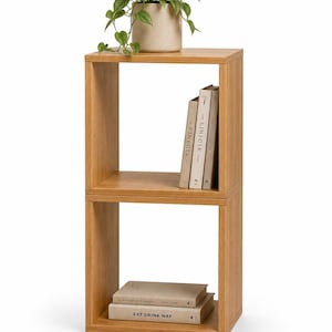 May include: A light brown wooden two-tier shelving unit with a square frame. The top shelf holds a beige pot with a green plant and three books titled Kinfolk. The bottom shelf contains two stacked books, with the bottom one labelled Eat Drink Nap.