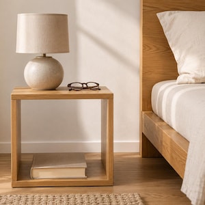 May include: A square light wood nightstand featuring an open shelf design. On top sits a round beige ceramic table lamp with a fabric shade and a pair of dark-rimmed reading glasses. A single closed book rests on the bottom shelf of the nightstand.