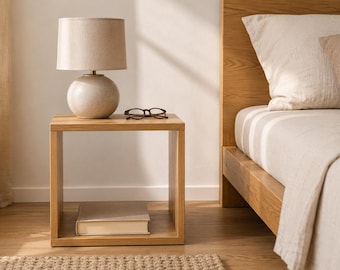 May include: A square light wood bedside table featuring an open shelf design. On top sits a round beige ceramic table lamp with a fabric shade and a pair of dark-rimmed reading glasses. A single closed book rests on the bottom shelf of the bedside table.