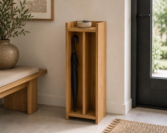 May include: A tall, light-toned wooden umbrella stand featuring a vertical divider that creates two narrow storage compartments. The top surface holds a small, round, light-colored stone bowl. A black umbrella is stored in the left section.