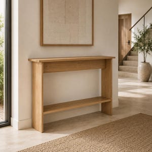 Peut inclure: Une console en bois brun clair dotée d'un plateau rectangulaire, de deux panneaux latéraux verticaux et d'une étagère inférieure. Ce meuble présente une finition en grain de bois naturel lisse et une structure linéaire sans aucune ornementation.
