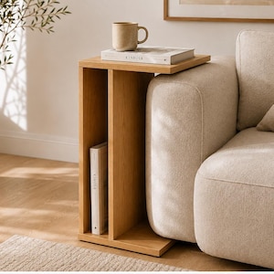 Puede incluir: Una mesa auxiliar en forma de C de madera de roble claro con un compartimento de almacenamiento vertical. La superficie superior plana sostiene una taza de cerámica beige y un libro titulado KINFOLK, con otro libro KINFOLK guardado verticalmente.