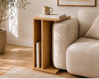 May include: A light oak wood C-shaped side table with a vertical storage compartment. The table features a flat top surface holding a beige ceramic mug and a book titled KINFOLK, with another KINFOLK book stored vertically in the lower section.