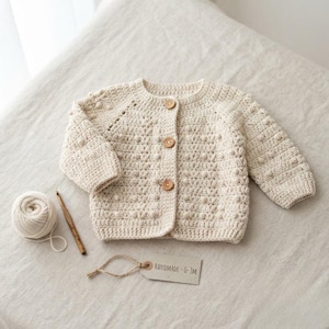 Könnte beinhalten: Eine cremefarbene, gehäkelte Langarmstrickjacke mit strukturiertem Noppenmuster und drei runden Holzknöpfen auf der Vorderseite. Ein Knäuel passendes Garn, eine Häkelnadel aus Holz und ein Etikett mit der Aufschrift "HANDMADE 0-3M" liegen bei.