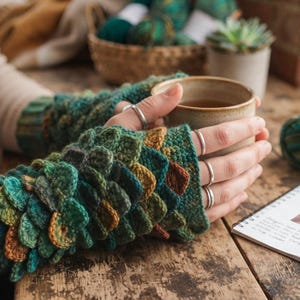 May include: Fingerless gloves featuring a textured, layered crochet pattern in variegated shades of forest green, teal, and brown. The gloves have a ribbed cuff and a scale-like design. A small notebook nearby displays the text Dragon Scales Crochet Gloves.