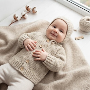May include: A beige knitted long-sleeve cardigan with a matching bonnet, featuring a textured stitch pattern and four small wooden buttons down the front. The garment is crafted with ribbed cuffs and a ribbed hem, providing a consistent look.