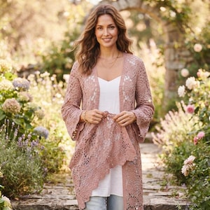 Op de afbeelding: Een oudroze vest met open voorzijde, voorzien van een ingewikkeld bloemenkantpatroon met lange, wijde mouwen en een geschulpte zoom, gedragen over een eenvoudig wit topje en een blauwe spijkerbroek voor een gelaagde look met textuur.